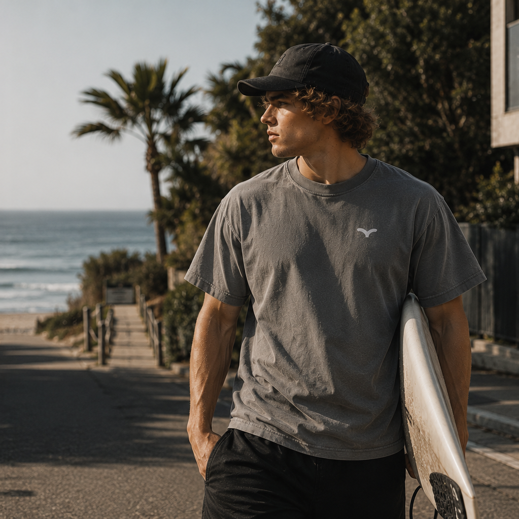 Person iført grå Endless Hope T-shirt med lille reflekslogo på brystet. T-shirten er fotograferet ved stranden med et surfboard under armen og understreger en afslappet livsstil tæt på havet.