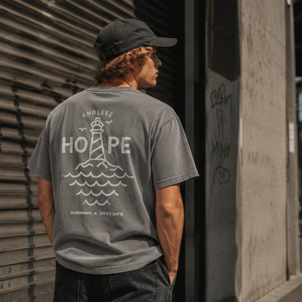 Person iført grå Endless Hope T-shirt fra Safe & Shine x Hjemhavn. Det store fyrtårnsmotiv på ryggen symboliserer håb, retning og frihed og er inspireret af livet ved kysten.
