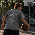 Person spiller basketball iført grå Endless Hope T-shirt fra Safe & Shine x Hjemhavn. Det store fyrtårnsmotiv på ryggen symboliserer håb, styrke og retning i bevægelse.