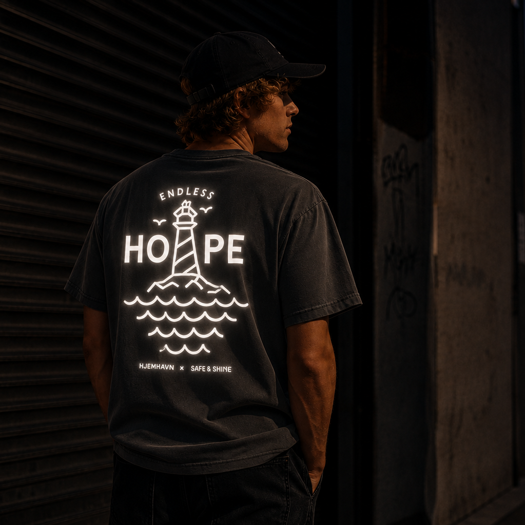 Person iført mørkegrå Endless Hope T-shirt fra Safe & Shine x Hjemhavn i aftenlys. Det store refleksprint med fyrtårn og bølger på ryggen lyser kraftigt i mørket.