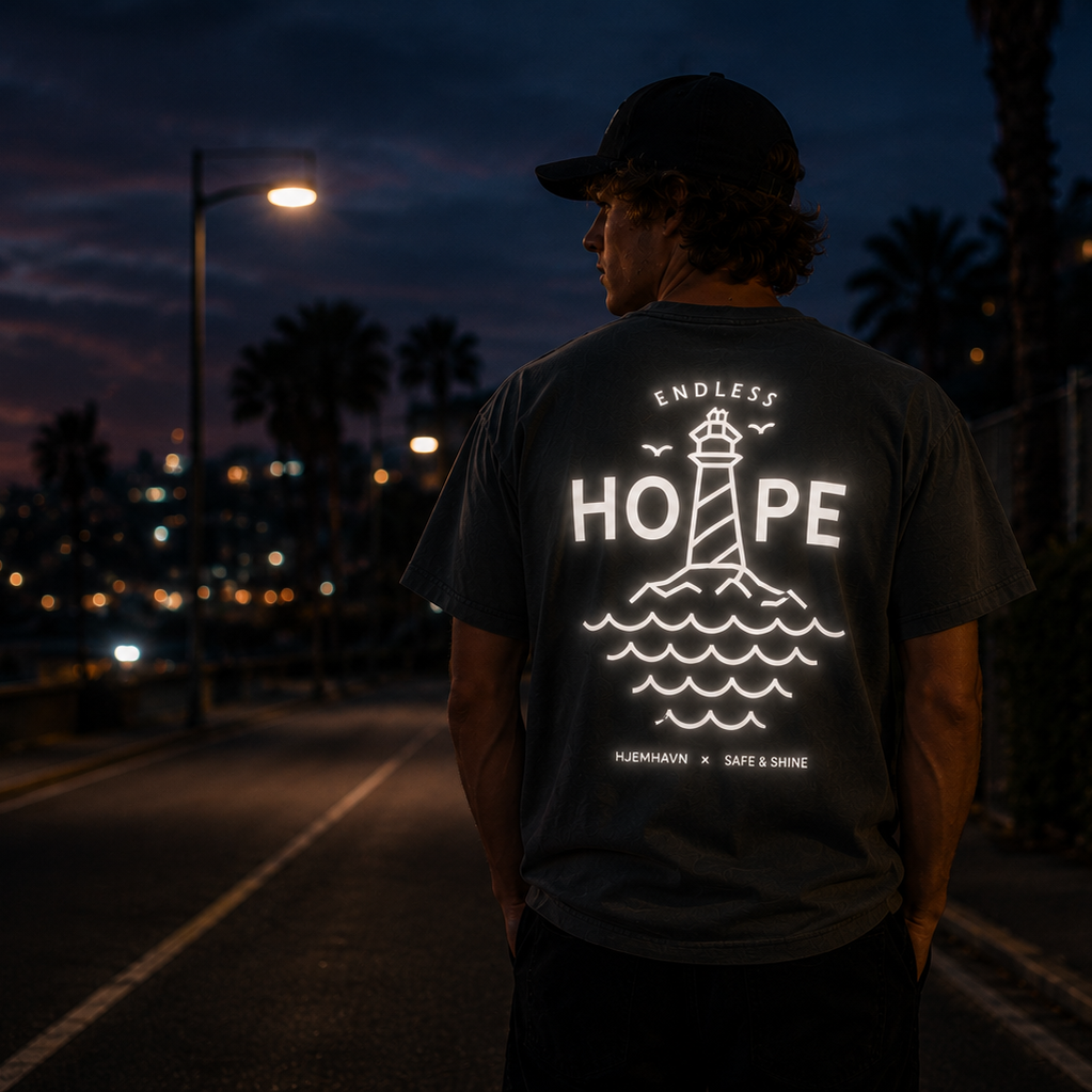 Person iført mørkegrå Endless Hope T-shirt fra Safe & Shine x Hjemhavn på en gade i aftentimerne. Det store refleksprint med fyrtårn og bølger på ryggen lyser kraftigt op i mørket og øger synligheden.