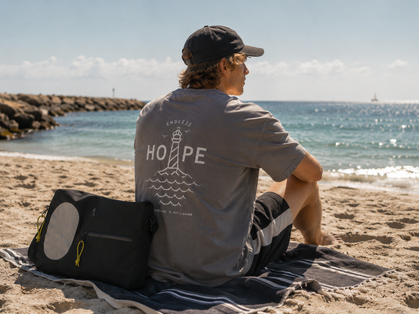 Mand på stranden iført grå ENDLESS HOPE T-Shirt fra HJEMHAVN x Safe & Shine med fyrtårnsmotiv på ryggen og Jupiter crossbody taske ved siden af.