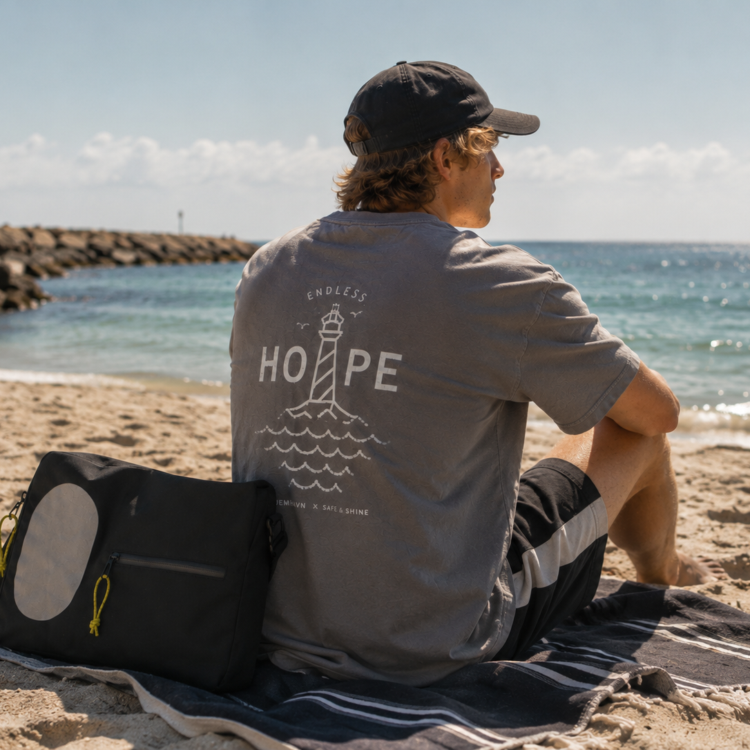 Person sidder ved stranden iført grå Endless Hope T-shirt fra Safe & Shine x Hjemhavn. Det store fyrtårnsmotiv på ryggen ses sammen med en sort refleks-computertaske med refleksdetaljer.