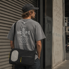 Person iført grå Endless Hope T-shirt fra Safe & Shine x Hjemhavn og sort refleks-computertaske. Det store fyrtårnsmotiv på ryggen kombineres med funktionelle refleksdetaljer til en aktiv hverdag.