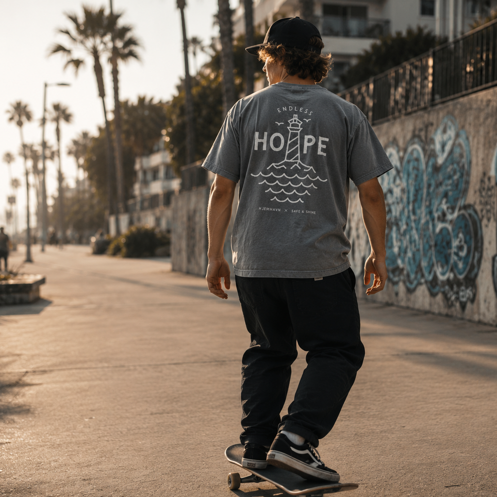 Person på skateboard iført grå Endless Hope T-shirt fra Safe & Shine x Hjemhavn. Det store fyrtårnsmotiv på ryggen symboliserer håb, retning og frihed i hverdagen.