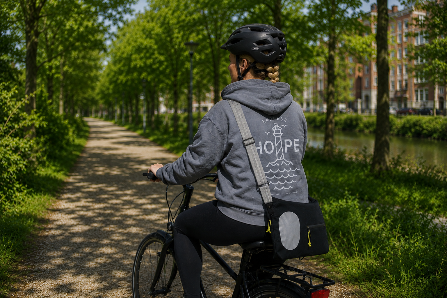 Kvinde på cykel iført ENDLESS HOPE hoodie og Safe & Shine Jupiter crossbody reflekstaske på grøn cykelsti.