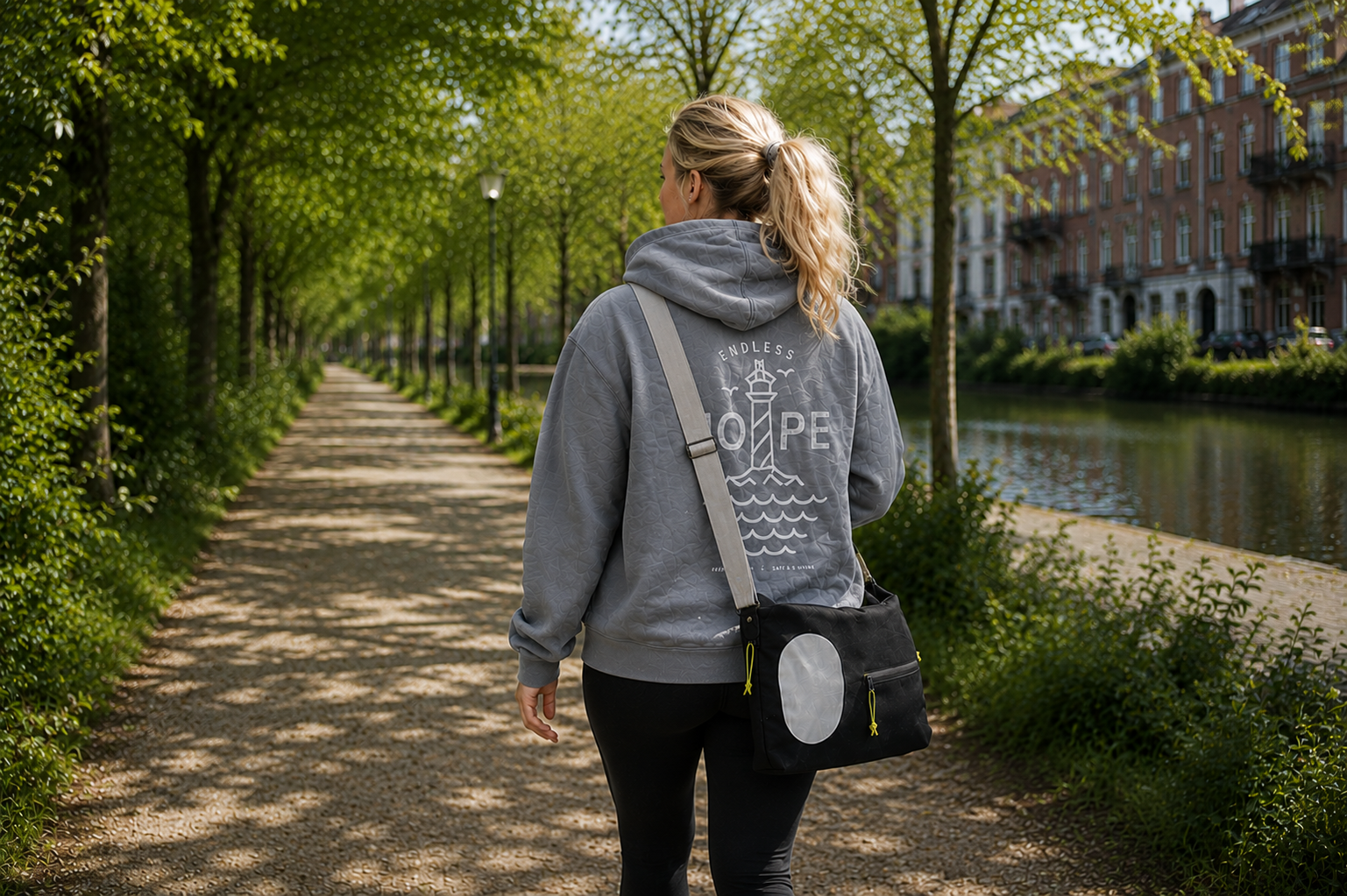 Kvinde på gåtur iført ENDLESS HOPE hoodie og Safe & Shine Jupiter crossbody reflekstaske på grøn cykelsti.