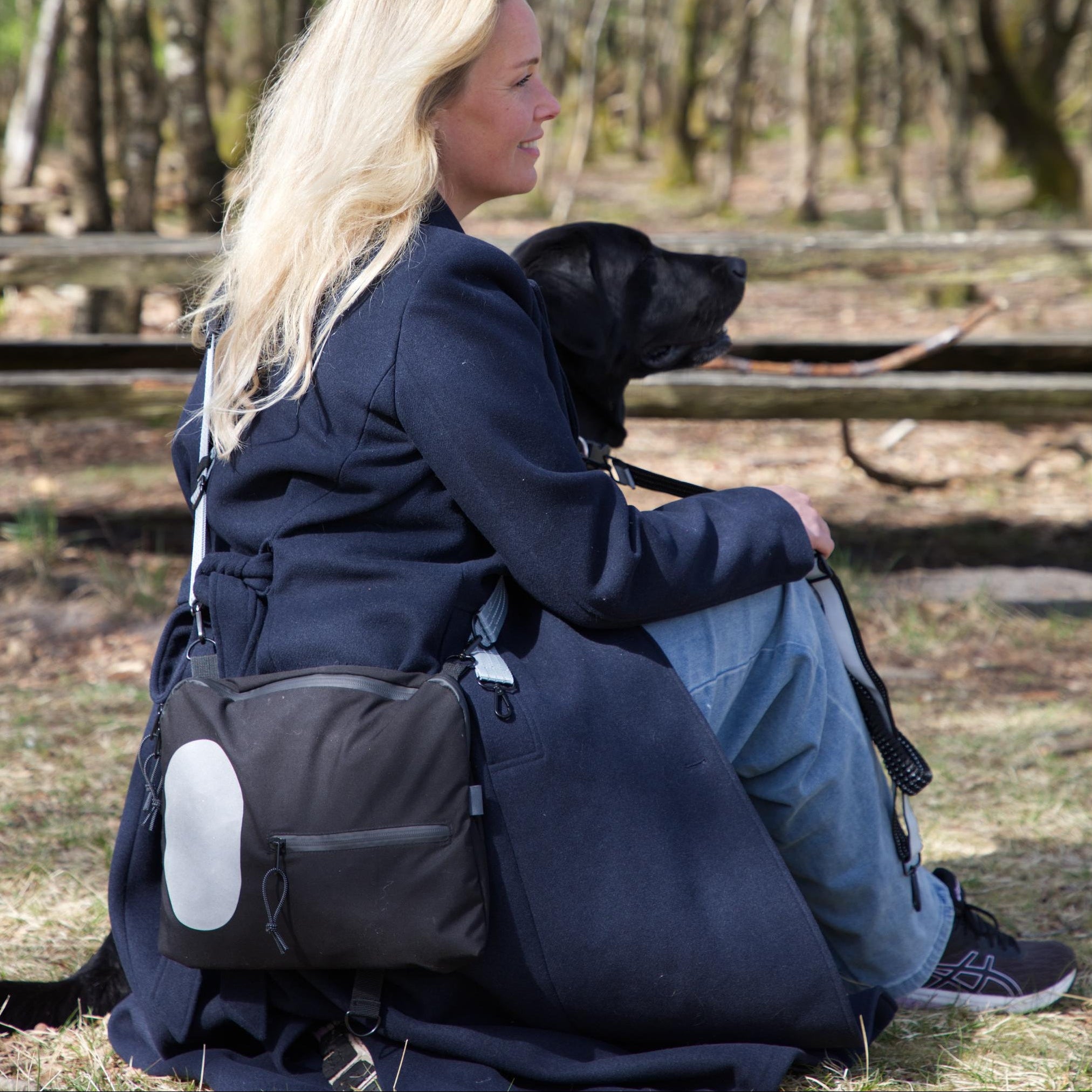 Kvinde med hund i naturen bærer sort skuldertaske med refleksdetalje fra Safe & Shine – synlighed til hverdag og gåture