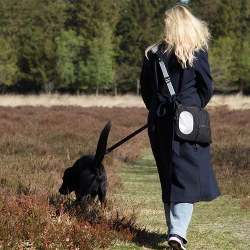 Kvinde går tur med hund i naturen og bærer sort skuldertaske med refleks fra Safe & Shine – synlighed til hverdag og bevægelse