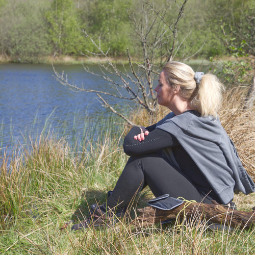 Kvinde sidder ved sø i naturen iført grå hoodie fra Safe & Shine – refleks og komfort som en naturlig del af hverdagen