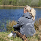 Kvinde sidder ved sø iført grå HOPE hoodie med refleksprint fra Hjemhavn x Safe & Shine – synlighed inspireret af natur og hverdagsliv