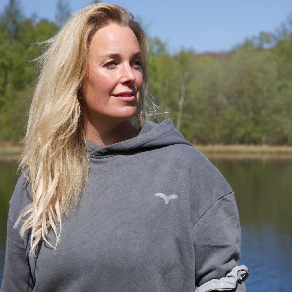 Kvinde ved sø iført grå hoodie med reflekslogo fra Safe & Shine – synlighed som en naturlig del af hverdagen