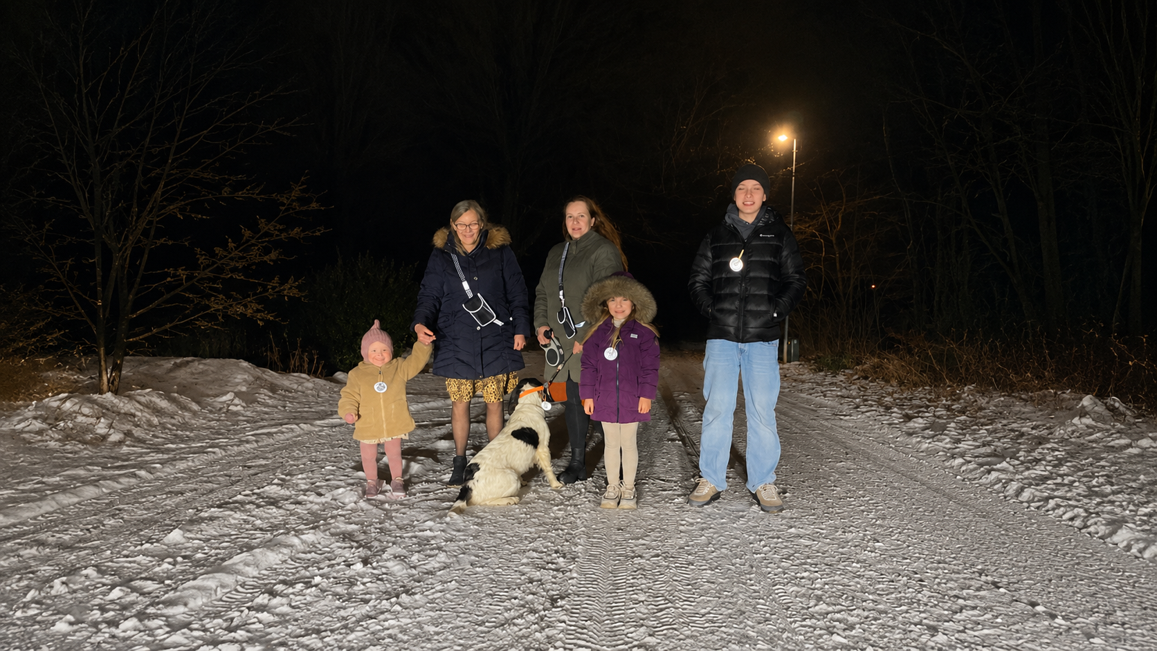 Familie med børn og hund på aftengåtur i sneen iført Safe & Shine refleksprodukter, herunder refleksstropper og refleksvedhæng, der øger synligheden i mørket.